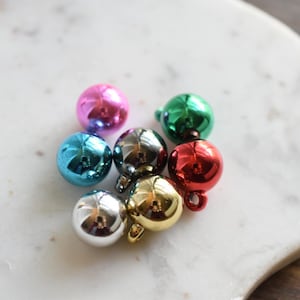 Puede incluir: Una colección de pequeños adornos metálicos redondos en varios colores, incluyendo rosa, azul, plateado, verde, rojo y dorado. Cada adorno tiene un pequeño lazo en la parte superior. Los adornos están dispuestos sobre una superficie de mármol blanco.