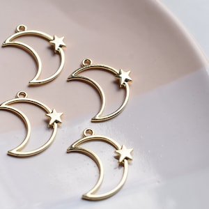 May include: Four gold metal crescent moon charms with a small star inside each moon. The charms are on a light pink and white surface.