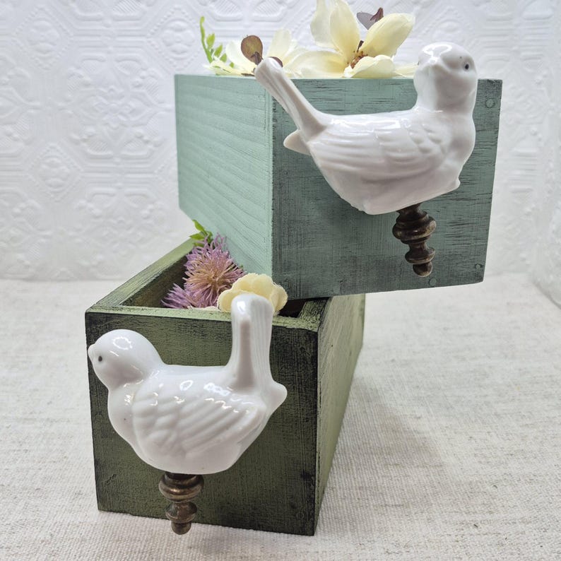 May include: Two wooden boxes with white porcelain bird drawer pulls. The top box is painted a light blue-green colour and the bottom box is painted a dark green colour. Both boxes have dried flowers inside.