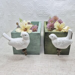 May include: Two wooden planter boxes with bird-shaped handles. The boxes are painted in a light green and dark green colour. The boxes are filled with dried flowers.