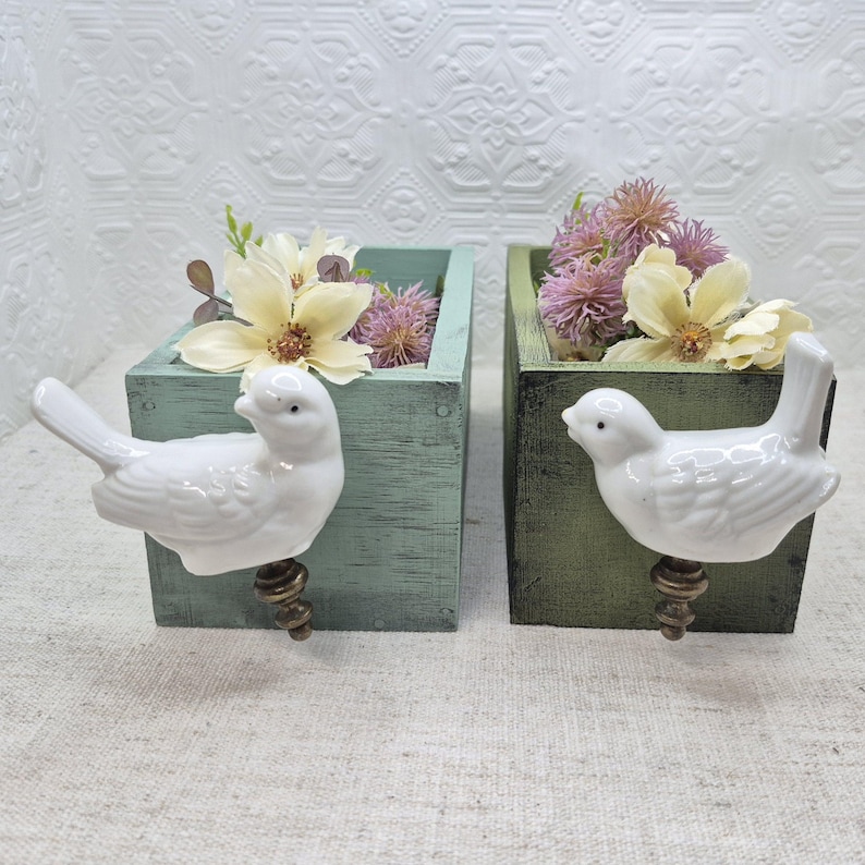 May include: Two wooden planter boxes with bird-shaped handles. The boxes are painted in a light green and dark green colour. The boxes are filled with dried flowers.