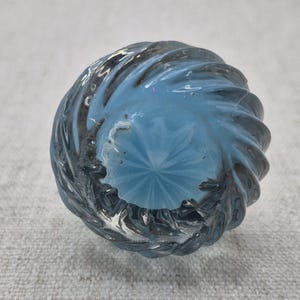 May include: A clear glass bottle stopper with a spiral design. The stopper has a light blue interior and a textured, swirled exterior. The bottom of the stopper has a starburst pattern.