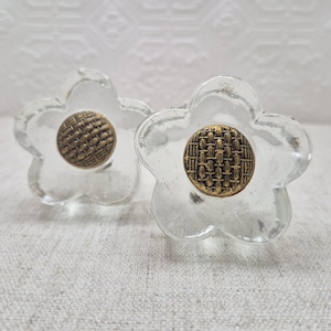 May include: Two clear glass flower-shaped cabinet knobs with a woven brass-colored center. The knobs have a vintage aesthetic and are suitable for furniture restoration or adding a decorative touch to drawers and cabinets.