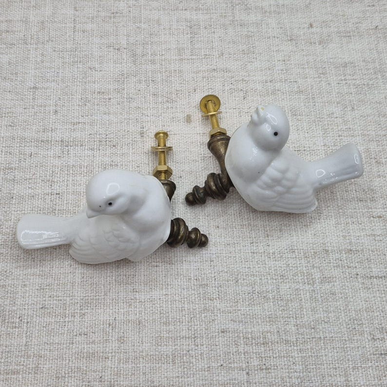 May include: Two white porcelain bird drawer pulls with gold metal hardware. The birds are facing to the right and have a detailed design.