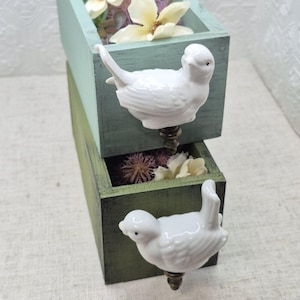 May include: Two wooden boxes with bird-shaped drawer pulls. The top box is painted a light blue-green colour and the bottom box is painted a dark green colour. Both boxes have dried flowers inside.