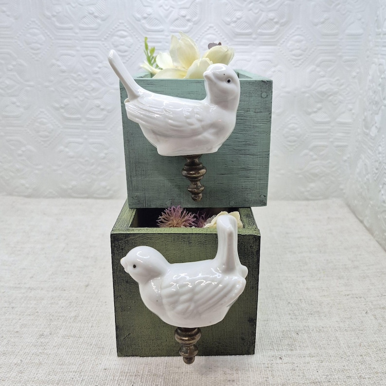 May include: Two wooden boxes, one painted green and the other blue, stacked on top of each other. Each box has a white porcelain bird knob with a brown metal stem.