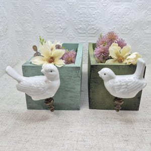 May include: Two wooden planter boxes with bird-shaped handles. The boxes are painted in a light green and dark green color. The boxes are filled with dried flowers.