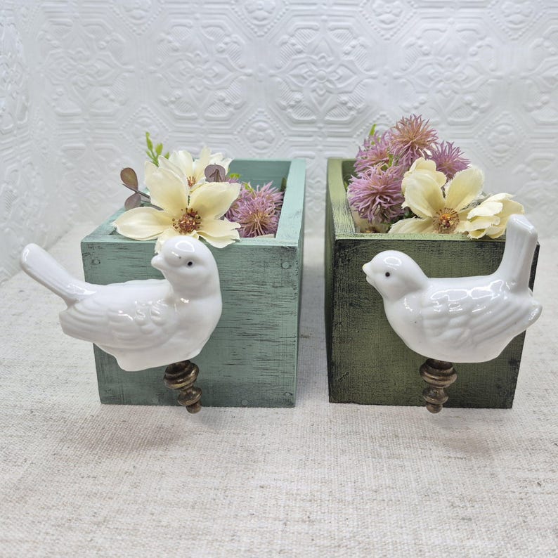 May include: Two wooden planter boxes with bird-shaped handles. The boxes are painted in a light green and dark green colour. The boxes are filled with dried flowers.