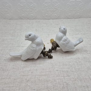 May include: Two white porcelain bird drawer pulls with gold metal accents. The birds are facing different directions and have detailed wings.