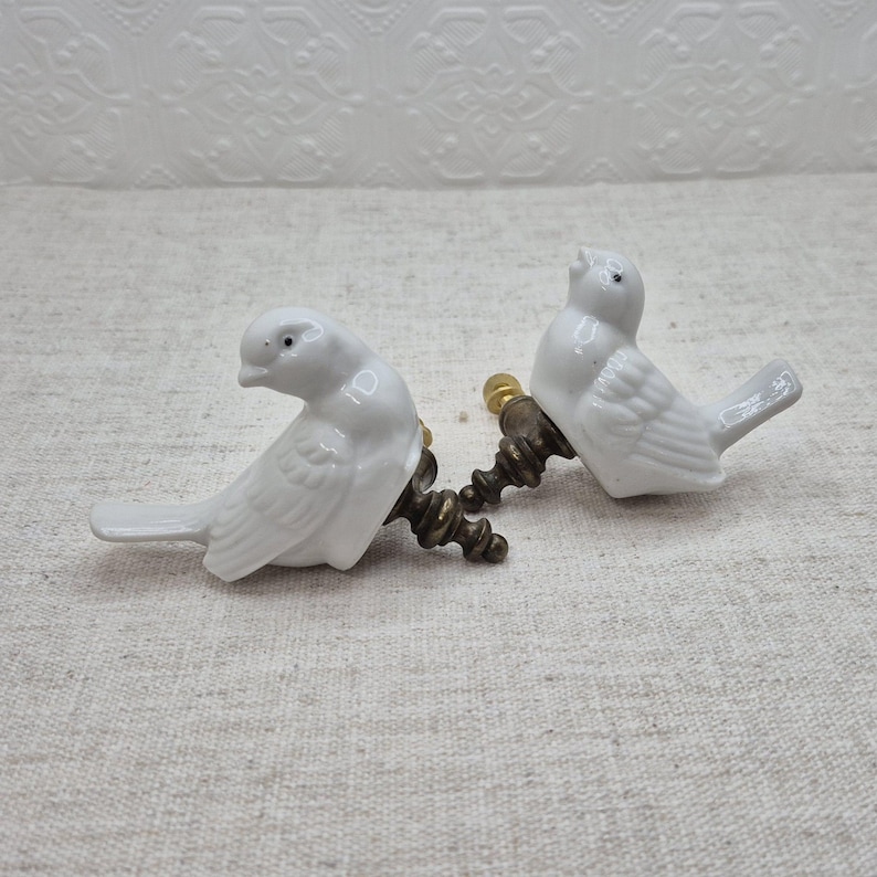 May include: Two white porcelain bird drawer pulls with gold metal accents. The birds are facing different directions and have detailed wings.