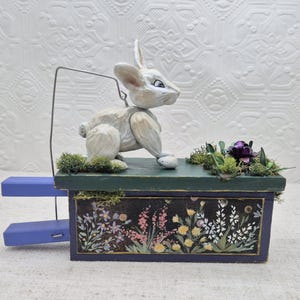 May include: A wooden rabbit puppet with a white and gray painted finish sits on a green and blue painted box. The box has a floral design and a small amount of moss and greenery on the top.
