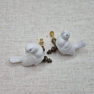 May include: Two white ceramic bird drawer pulls with gold metal hardware. The birds are facing to the right and have a detailed design.