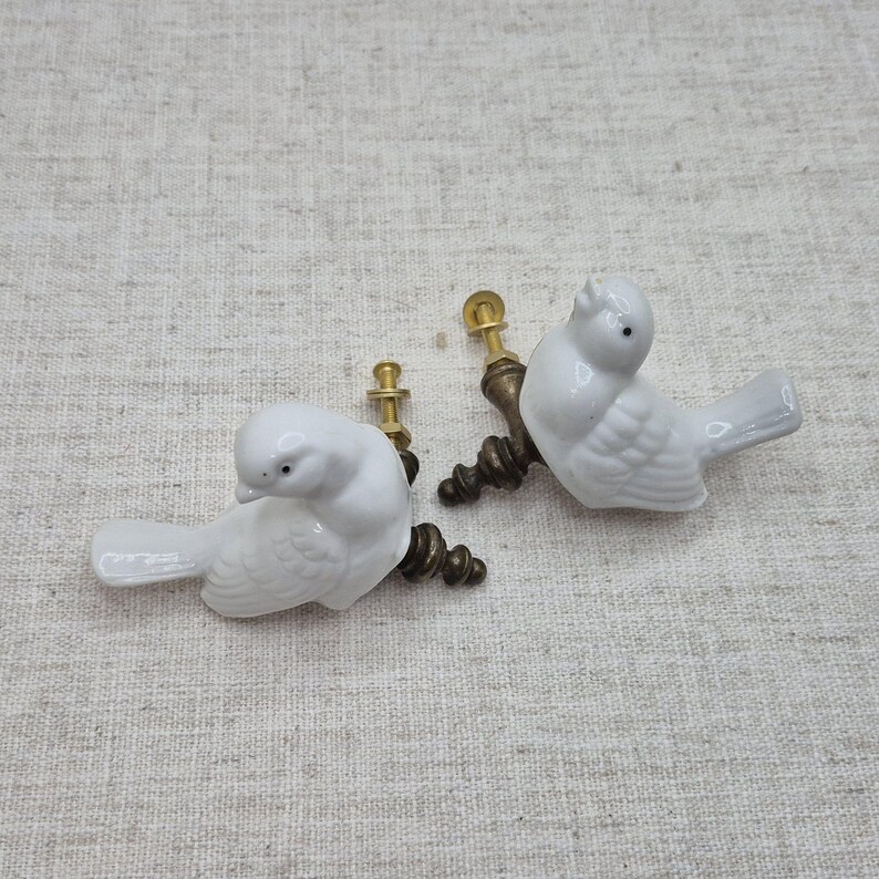 May include: Two white ceramic bird drawer pulls with gold metal hardware. The birds are facing to the right and have a detailed design.