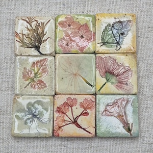 May include: A set of nine square ceramic tiles arranged in a grid, each featuring a unique botanical print. The designs include pressed flowers, leaves, and a dandelion seed head in shades of pink, green, brown, and cream on a textured surface.