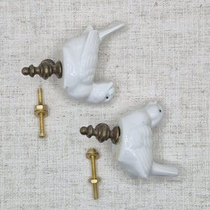 May include: Two white ceramic bird finials with gold metal accents and screws. The finials are in the shape of birds with their wings spread out. The screws are used to attach the finials to a surface.