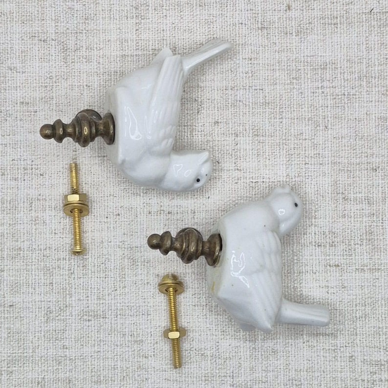 May include: Two white ceramic bird finials with gold metal accents and screws. The finials are in the shape of birds with their wings spread out. The screws are used to attach the finials to a surface.