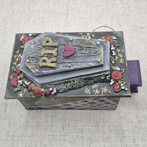 May include: A decorative box with a coffin-shaped lid, featuring the letters "RIP" and a heart. The box is adorned with red roses and floral accents, with a purple pull-out drawer. The overall design has a gothic theme.