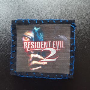May include: A square patch with the Resident Evil 2 logo. The image shows a close-up of a character's face with the game title in red and white. The patch is framed with dark blue fabric and stitched with blue thread.