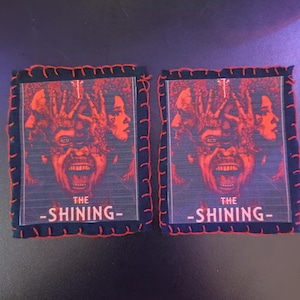 May include: Two square fabric patches featuring a red and black graphic from the movie "The Shining." The design includes a screaming face and the film's title in white text. The edges are stitched with red thread.