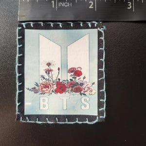 Może przedstawiać: Kwadratowa naszywka z jasnoniebieskim tłem z kwiatowym wzorem i białymi literami "BTS". Naszywka jest obramowana czarną tkaniną i jasnoniebieskimi szwami. Nad naszywką umieszczono linijkę o długości ok. 7,6 cm.