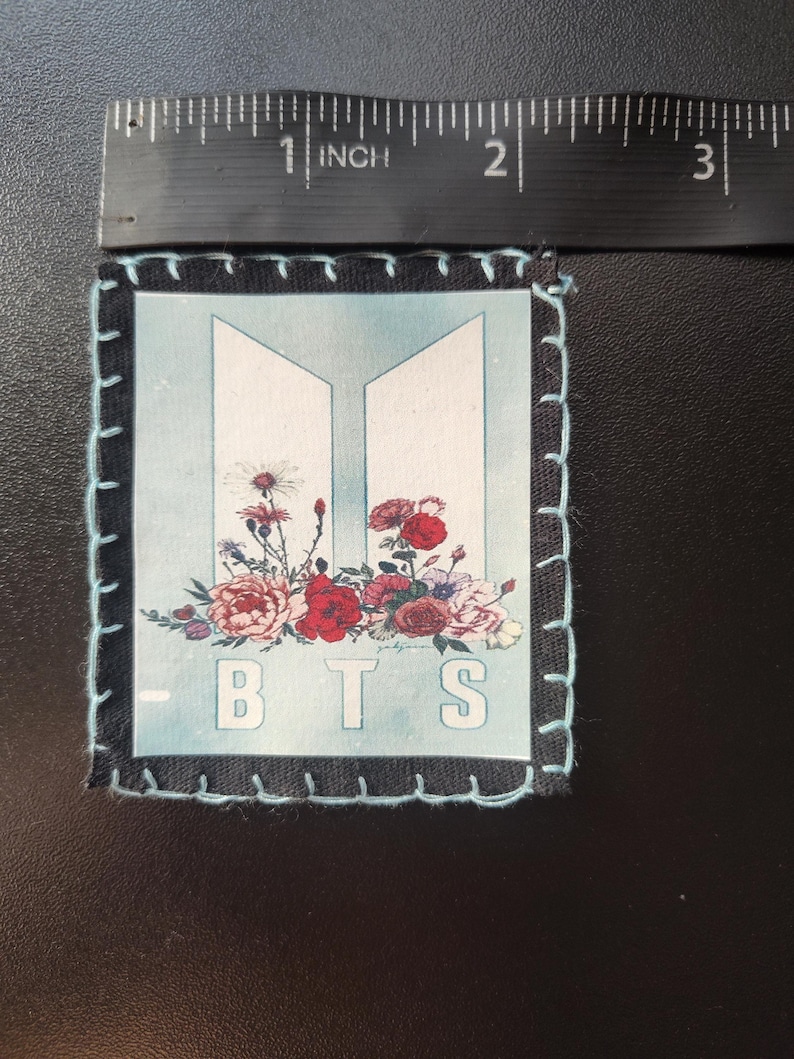 Może przedstawiać: Kwadratowa naszywka z jasnoniebieskim tłem z kwiatowym wzorem i białymi literami "BTS". Naszywka jest obramowana czarną tkaniną i jasnoniebieskimi szwami. Nad naszywką umieszczono linijkę o długości ok. 7,6 cm.