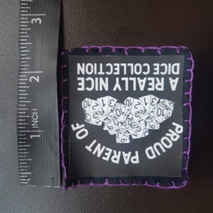 May include: Black square patch with purple stitching around the edges. The patch has the text "A REALLY NICE DICE COLLECTION" and "PROUD PARENT OF" in white. A cluster of white dice is in the center. The patch is approximately 2 inches wide.