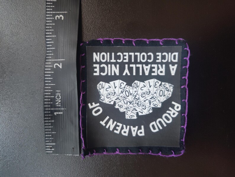 May include: Black square patch with purple stitching around the edges. The patch has the text "A REALLY NICE DICE COLLECTION" and "PROUD PARENT OF" in white. A cluster of white dice is in the center. The patch is approximately 2 inches wide.