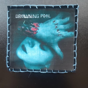 May include: Square patch with a blue and white stitched border. The image features a person's face and hand with the word "SINNER" tattooed on the fingers. The text "DROWNING POOL" is at the top.