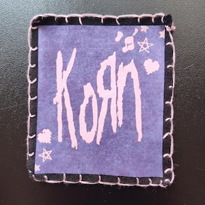 May include: Square patch with the band name "Korn" in white, stylized lettering on a purple background. The patch has a black border with pink stitching and small star and musical note graphics.