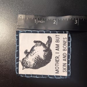 May include: A rectangular patch with a black and white illustration of a cat lying on its back. The patch is approximately 2 inches wide and has the text "MOTHER, I AM BUT SKIN AND BONES". The patch is stitched with light blue thread.