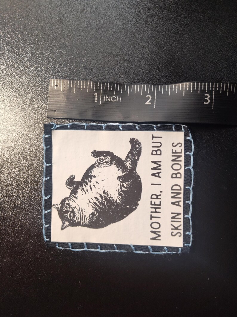 May include: A rectangular patch with a black and white illustration of a cat lying on its back. The patch is approximately 2 inches wide and has the text "MOTHER, I AM BUT SKIN AND BONES". The patch is stitched with light blue thread.