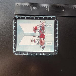 Może przedstawiać: Prostokątna naszywka z jasnoniebieskim tłem i białymi literami "BTS". Naszywka jest ozdobiona kompozycją kwiatową z czerwonych, r&oacute;żowych i białych kwiat&oacute;w. Naszywka ma około 6,35 cm długości.