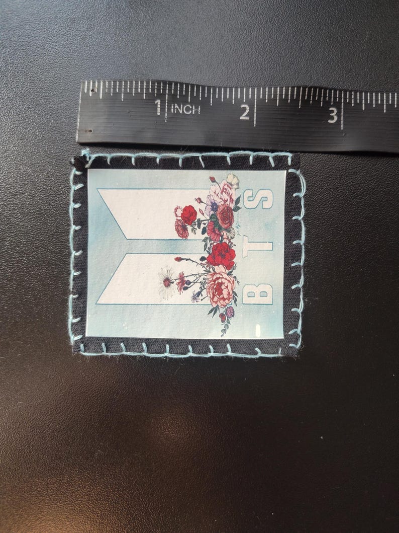 Może przedstawiać: Prostokątna naszywka z jasnoniebieskim tłem i białymi literami "BTS". Naszywka jest ozdobiona kompozycją kwiatową z czerwonych, r&oacute;żowych i białych kwiat&oacute;w. Naszywka ma około 6,35 cm długości.