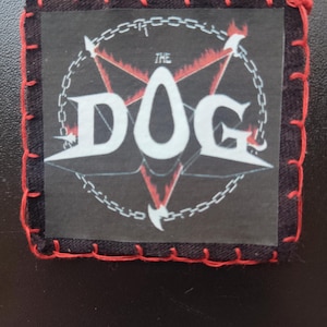 May include: Square black patch with red stitching around the edges. The patch features a white pentagram design with the words "THE DOG" in white, encircled by a chain. Red flames emanate from the pentagram.