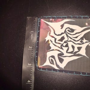 K&ouml;nnte beinhalten: Quadratischer Aufn&auml;her mit einem wei&szlig;en, abstrakten Design auf transparentem Hintergrund. Der Aufn&auml;her ist etwa 7,6 cm lang und breit, mit einem t&uuml;rkis gen&auml;hten Rand. Ein Lineal liegt neben dem Aufn&auml;her.