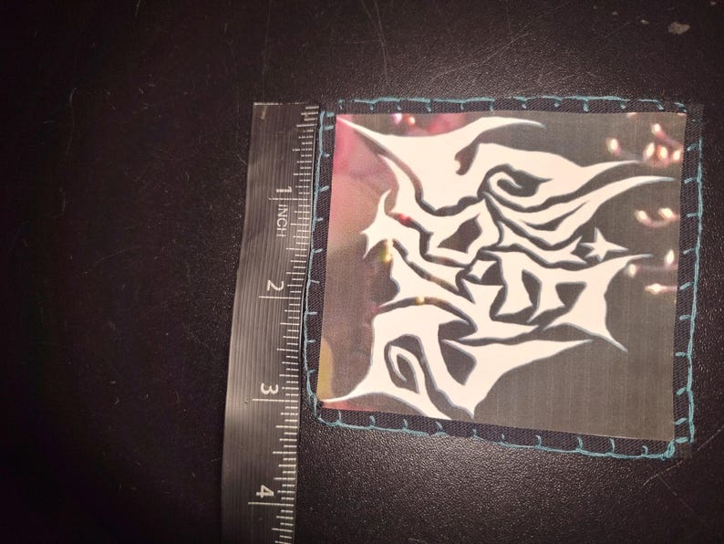 K&ouml;nnte beinhalten: Quadratischer Aufn&auml;her mit einem wei&szlig;en, abstrakten Design auf transparentem Hintergrund. Der Aufn&auml;her ist etwa 7,6 cm lang und breit, mit einem t&uuml;rkis gen&auml;hten Rand. Ein Lineal liegt neben dem Aufn&auml;her.