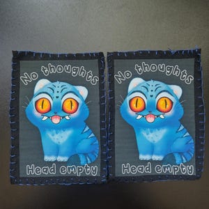 May include: Two square patches with a cartoon blue cat design. The cat has large orange eyes, a pink tongue, and the text "No thoughts" and "Head empty" in white. The patches have a black border with blue stitching.