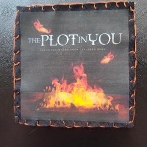 May include: Square patch featuring the text "THE PLOT IN YOU" above a fiery image. The patch has a black border with orange stitching.