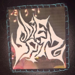 May include: A square patch with a white, stylized design on a transparent background. The design is framed by a black fabric border with teal stitching. The background behind the design is a blurred image with pink and yellow hues.