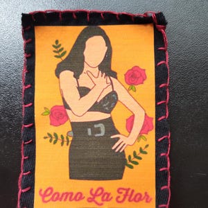 May include: A rectangular patch with a black border and red stitching. The patch features an illustration of a woman in a black outfit, with the text "Como La Flor" in pink. The background is orange with pink roses.