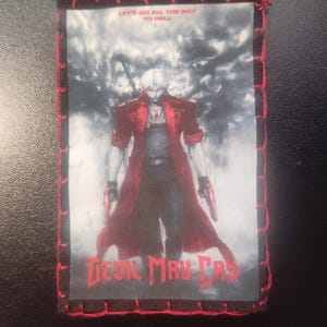 May include: A rectangular patch featuring a character from Devil May Cry, in a red coat, holding two guns. The patch has a red stitched border and the text "LET'S GO ALL THE WAY TO HELL" and "DEVIL MAY CRY".