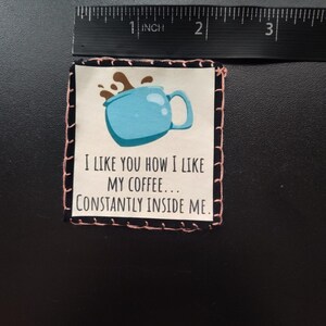 K&ouml;nnte beinhalten: Ein quadratischer Stoffflicken mit einer hellblauen Kaffeetassen-Illustration und dem Text "I LIKE YOU HOW I LIKE MY COFFEE... CONSTANTLY INSIDE ME." Der Flicken ist etwa 5 cm breit und hat einen schwarz-rosa gen&auml;hten Rand.