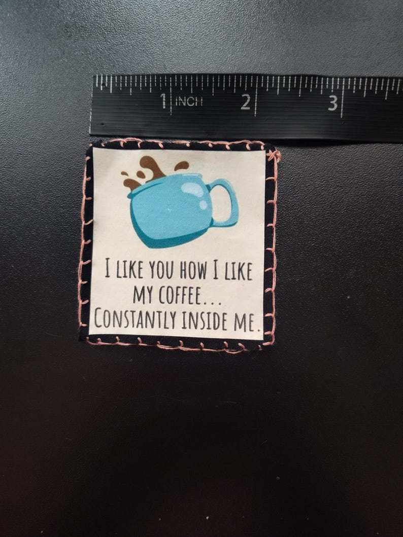 K&ouml;nnte beinhalten: Ein quadratischer Stoffflicken mit einer hellblauen Kaffeetassen-Illustration und dem Text "I LIKE YOU HOW I LIKE MY COFFEE... CONSTANTLY INSIDE ME." Der Flicken ist etwa 5 cm breit und hat einen schwarz-rosa gen&auml;hten Rand.