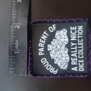 May include: A black patch with purple stitching around the edges. The patch has the text "PROUD PARENT OF A REALLY NICE DICE COLLECTION" in white. A cluster of dice is in the center. The patch is approximately 2 inches long.