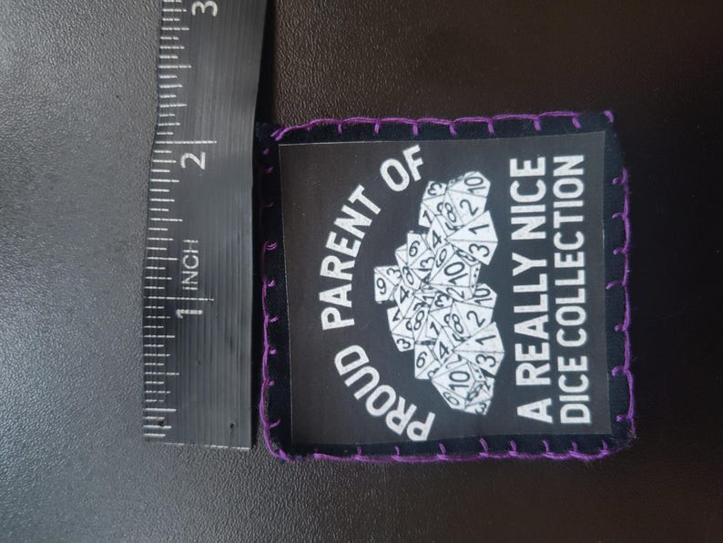 May include: A black patch with purple stitching around the edges. The patch has the text "PROUD PARENT OF A REALLY NICE DICE COLLECTION" in white. A cluster of dice is in the center. The patch is approximately 2 inches long.