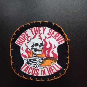 May include: Black patch with a white skull holding a taco, surrounded by red flames. The text reads "HOPE THEY SERVE TACOS IN HELL" in red. The patch has an orange stitched border.