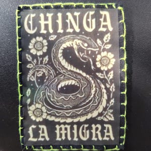 May include: Black patch with a snake illustration and the words "CHINGA LA MIGRA" in white. The patch has a black border with neon green stitching.