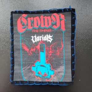 Peut inclure: Un écusson carré avec les mots "Crowng" en rouge, lettrage stylisé. En dessous, le texte "The Empire" et "Voriols" est visible. L'écusson présente un motif graphique bleu et rouge et est bordé de tissu noir et de coutures bleues.