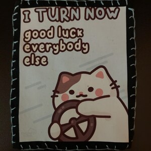May include: A rectangular patch with a black border and blue stitching. The patch features a cartoon cat driving a steering wheel, with the text "I TURN NOW good luck everybody else". The cat is white with brown spots and pink cheeks.