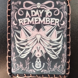 May include: Square patch with the text "A Day To Remember" in white and pink. The patch has a black border with pink stitching. The design includes a skull, wings, and other graphic elements.